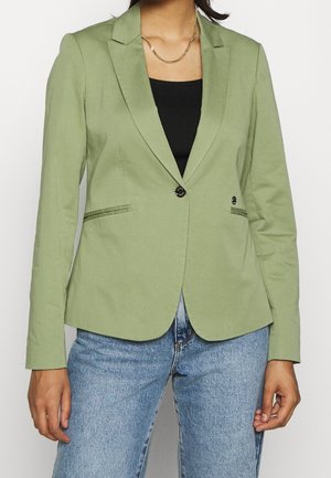 Vrouw draagt een lichtgroene enkelvoudige knoop blazer over een zwarte top met blauwe jeans, een gouden ketting zichtbaar bij de nek.
