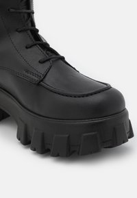Černá kožená bota s masivní podrážkou s drážkami a vyšitým detailem na špičce, s šněrováním.