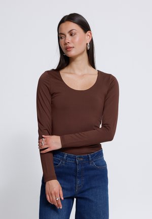 Vrouw met lang donker haar die zilveren creolen draagt, een bruin shirt met lange mouwen, blauwe jeans en meerdere ringen aan haar vingers.