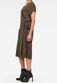 Vestido envolvente verde oliva con mangas cortas, bolsillos laterales y una cintura atada. Combinado con botas negras hasta el tobillo y un corte recto y elegante.