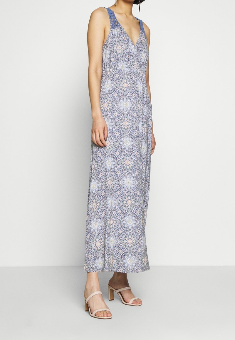 Robe maxi sans manches bleue avec un motif floral, dotée d'un décolleté en V et de délicats accents en dentelle sur les bretelles. Assortie à des sandales à talons blocs beiges.