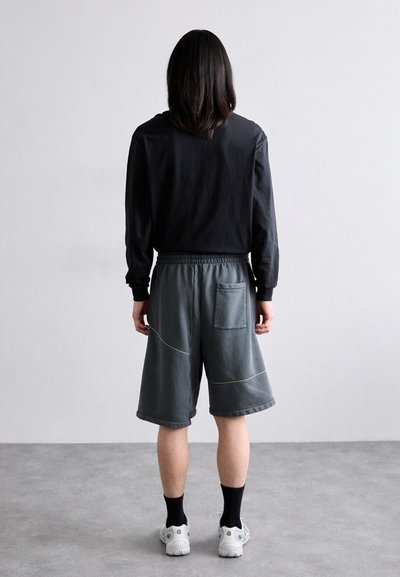 Locker sitzende graue Shorts mit gelben Nähten, elastischem Taillenbund und einer Gesäßtasche, kombiniert mit einem schwarzen Langarmshirt.