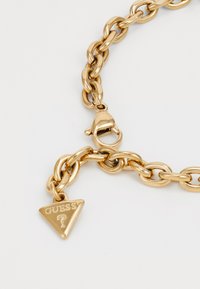 Lănțișor de gât din metal auriu, gros, cu un pandantiv triangular pe care este gravat "GUESS" și un semn de întrebare, prins cu un închizător.
