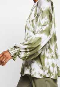 Osoba ubrana w zielono-białą bluzkę z długim rękawem w stylu tie-dye z rozciągniętymi mankietami, w połączeniu z oliwkowymi spodniami, idąca na tle jednolitym.