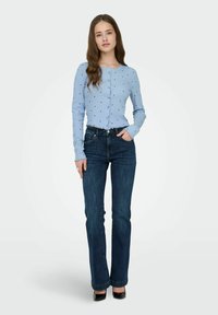 Cardigan bleu clair boutonné avec des points noirs, associé à un jean bleu foncé. Le cardigan possède des manches longues et des poignets festonnés. Talons hauts noirs.