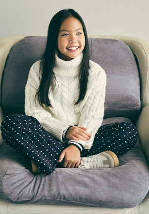 Ragazza sorridente con lunghi capelli scuri seduta a gambe incrociate su un cuscino grigio, indossa un maglione bianco a trecce, pantaloni blu navy con fantasia e sneakers bianche.