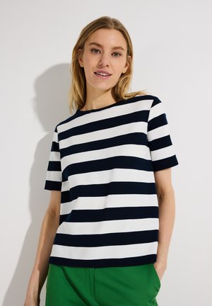 Junge Frau trägt ein schwarz-weiß gestreiftes T-Shirt mit kurzen Ärmeln und grüne Hose, steht vor einer schlichten weißen Wand.