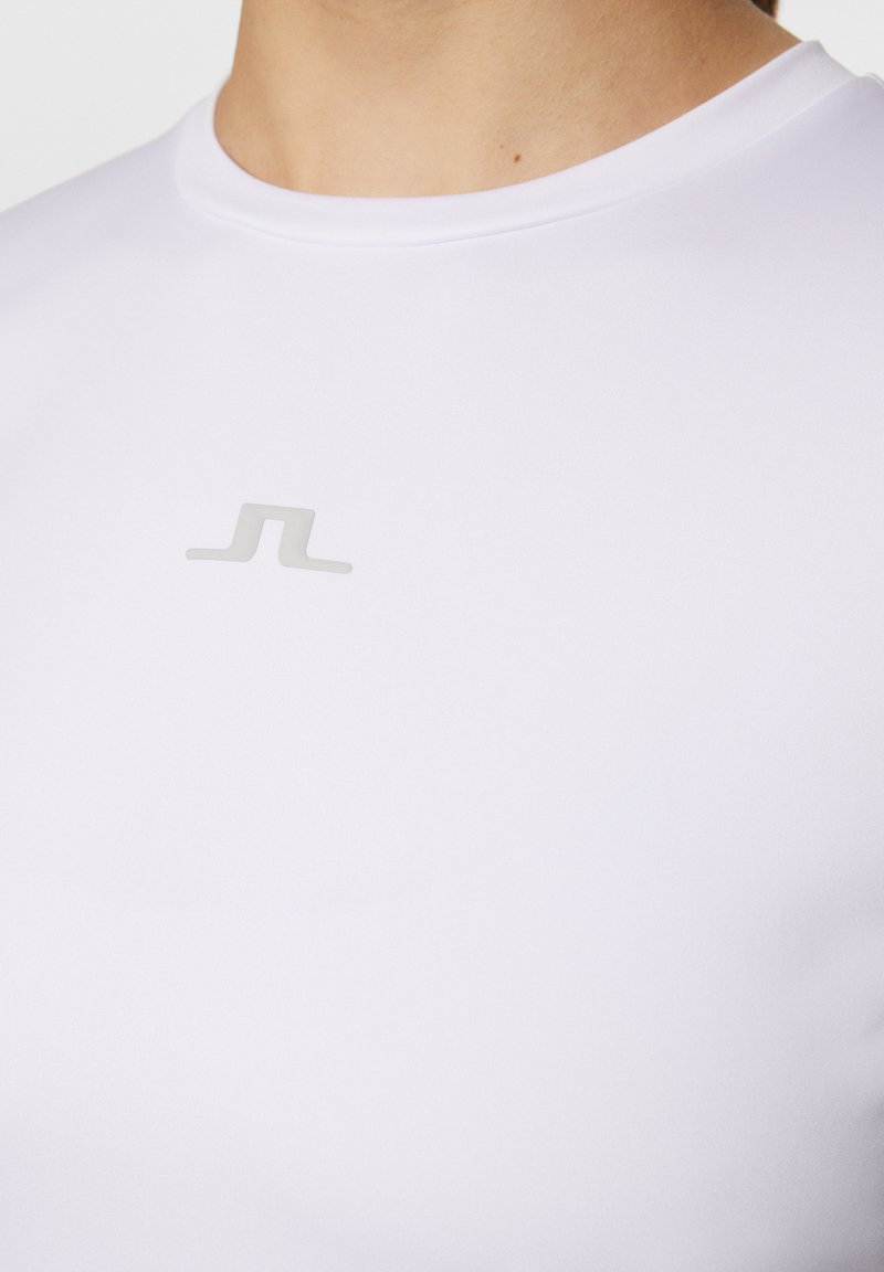 Maglietta sportiva bianca realizzata in un tessuto liscio e elastico. Presenta un logo grigio minimalista centrato sul petto. Collo rotondo.