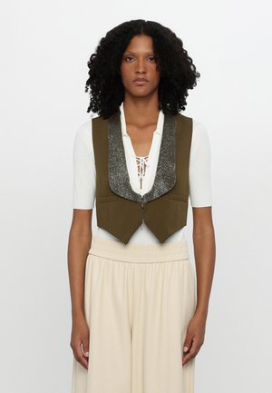 Olive green cropped vest with a glittery silver collar, features a zip closure and two front pockets, worn over a white top and beige pants.