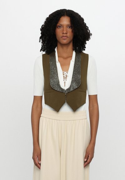 Veste courte vert olive avec un col argenté pailleté, dotée d'une fermeture éclair et de deux poches avant, portée par-dessus un top blanc et un pantalon beige.