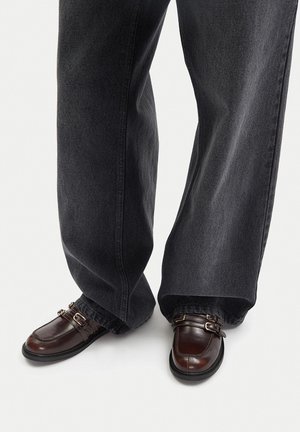 Tmavě šedé široké džíny kombinované s hnědými koženými botami s dvojitými přezkami a hladkým povrchem.