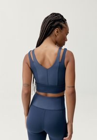 Tamsiai mėlynas sportinis liemenukas su prigludusia forma, dvigubomis petnešomis ir tekstūruotu audiniu. Turi V formos nugarą ir minimalias siūles paramai.