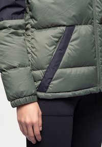 Polstret jakke med et olivengrønt rutemønster og marineblå detaljer, glidelås foran og ribbestrikkede mansjetter. Mørke leggings er delvis synlige.