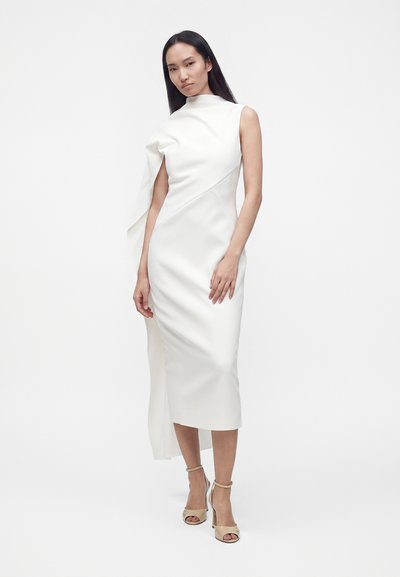 Femme aux longs cheveux foncés portant une robe midi blanche sans manches avec un tissu drapé asymétrique et des talons beiges à bride à la cheville.