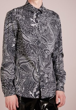 Mężczyzna nosi czarną, zapinaną na guziki koszulę z długim rękawem, ozdobioną misternymi białymi abstrakcyjnymi i organicznymi wzorami, w połączeniu z błyszczącymi czarnymi spodniami.