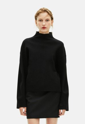 Vrouw in een zwart oversized trui met hoge hals en lange mouwen, gecombineerd met een zwarte mini rok, staande tegen een effen witte achtergrond.