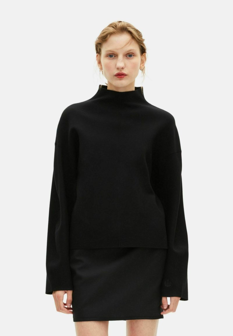 Donna che indossa un maglione oversized nero a collo alto con maniche lunghe e una minigonna nera, in piedi davanti a uno sfondo bianco semplice.