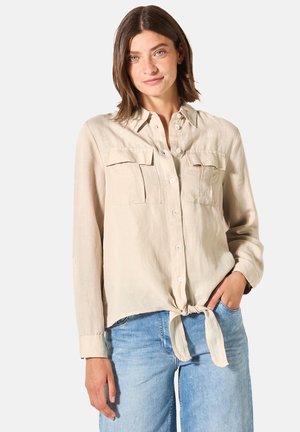 Chemise en lin beige clair avec un devant à boutons, deux poches poitrine et un ourlet noué. Manches longues avec poignets à boutons. Portée avec du denim.