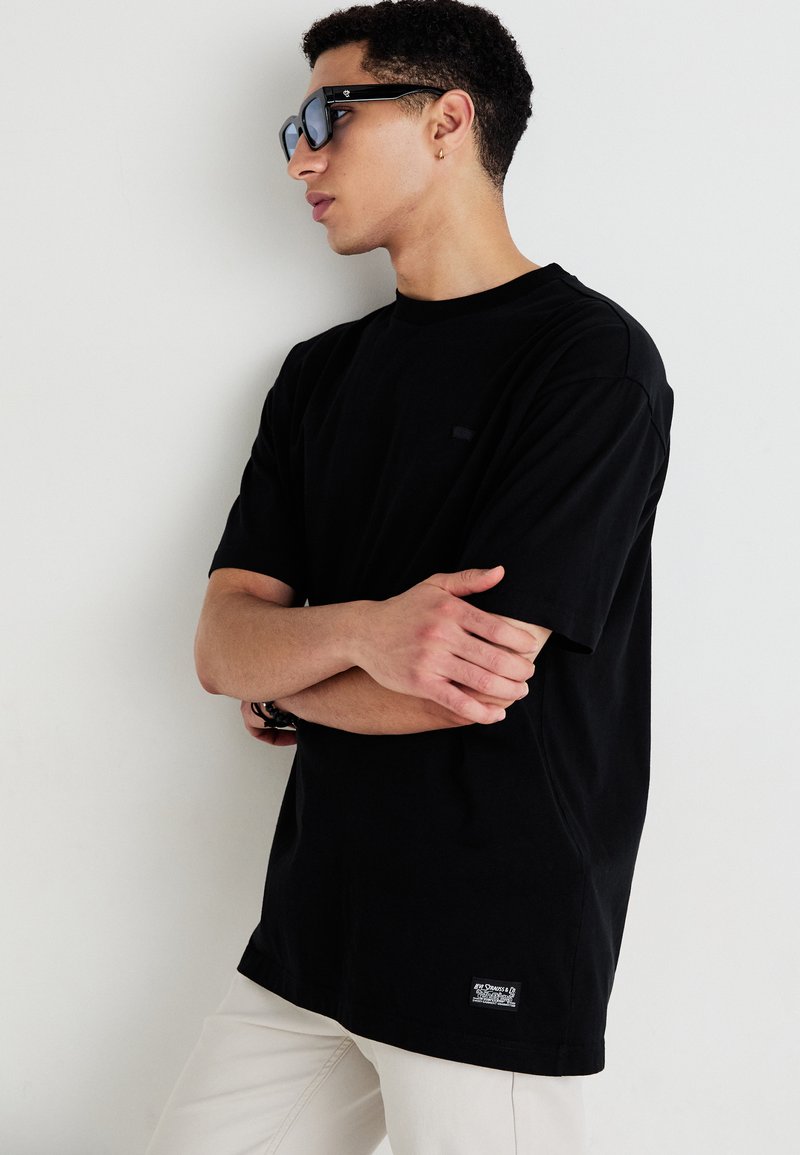 Czarny, krótki rękaw t-shirt z bawełny, z okrągłym dekoltem i małym detalem logo na klatce piersiowej. Biały metka na dolnym końcu. Noszony z okularami przeciwsłonecznymi.