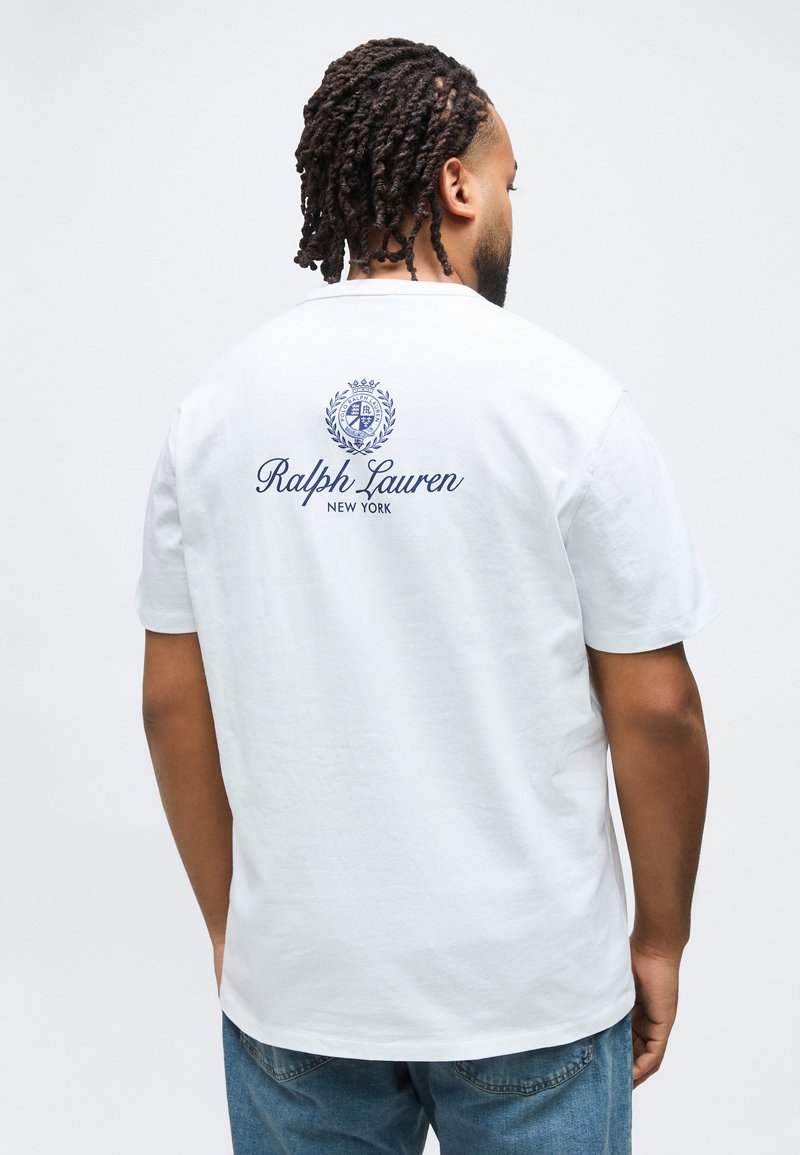 Άνδρας με πλεγμένα μαλλιά που φοράει λευκό t-shirt Ralph Lauren με μπλε λογότυπο και κείμενο στην πλάτη, συνδυασμένο με μπλε τζιν, γυρισμένος με την πλάτη.