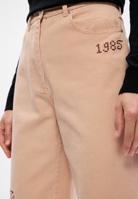 Μπεζ τζιν παντελόνι με χαλαρή εφαρμογή, πέντε τσέπες και κεντημένη λεπτομέρεια "1985" στο αριστερό μπούτι. Υφασμάτινη υφή.