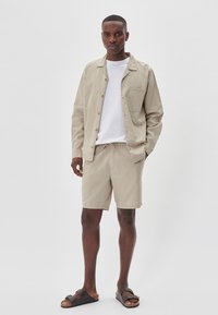 Beżowa bawełniana koszula z krótkimi rękawami z drewnianymi guzikami, zestawiona z beżowymi szortami i czarnymi sandałami na płaskiej podeszwie. Prosty, luźny fason i praktyczny design.