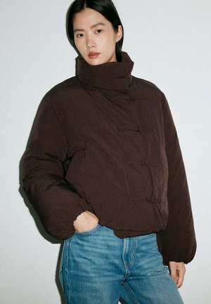 Mujer con cabello negro liso que lleva una chaqueta acolchada marrón con cuello alto y jeans azules, de pie con una mano en el bolsillo contra un fondo liso.