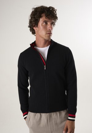 Zwarte gebreide zip-up jas met een textuurpatroon en rode accenten op de kraag en gestreepte manchetten. G gedragen over een wit T-shirt.