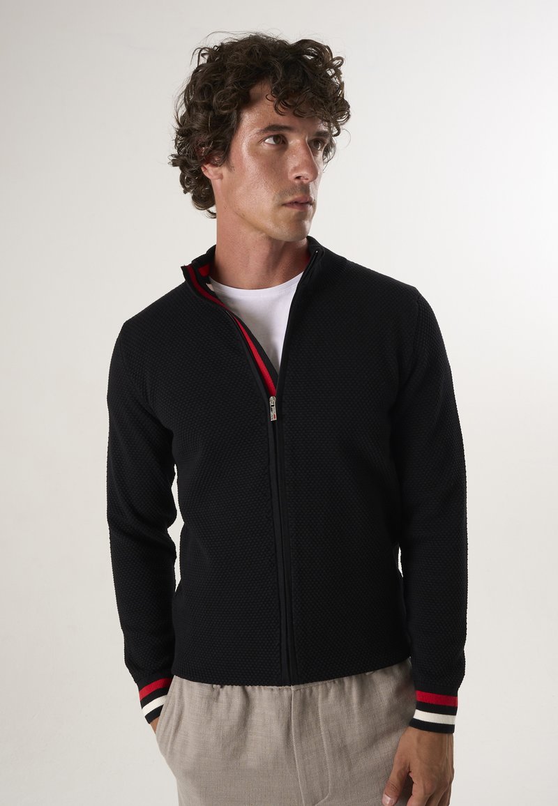 Zwarte gebreide zip-up jas met een textuurpatroon en rode accenten op de kraag en gestreepte manchetten. G gedragen over een wit T-shirt.