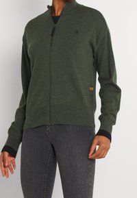 Donkergroene ritscardigan gemaakt van gestructureerde brei-stof, voorzien van een hoge kraag en contrasterende zwarte ribgebreide manchetten en zoom.