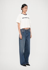 T-shirt bianca a maniche corte con scollatura navy e testo "MICHAEL KORS", abbinata a jeans in denim blu a gamba larga e scarpe nere a punta.