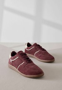Bordeaux-farbene Wildledersneaker mit cremefarbenen Akzenten, Schnürdesign und strukturiertem Gummisohle. Seitliche Streifen verleihen der Gesamtform zusätzliche Detail.