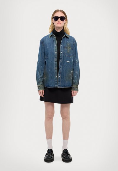 Jachetă din denim cu o nuanță ușor estompată de albastru, buzunar frontal și detalii cu stropi de vopsea, purtată peste un pullover negru cu guler înalt și o fustă mini.
