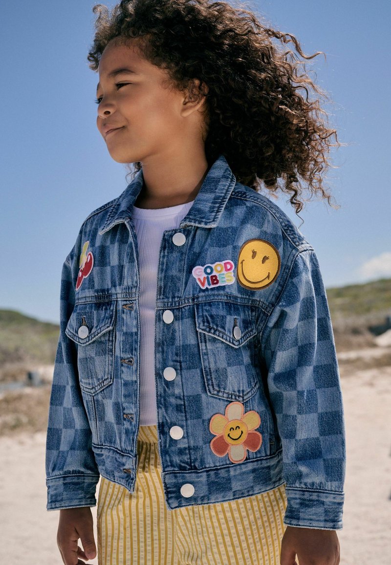 Barn med krøllet hår iført en blå ternet denimjakke med patches og gule stribede bukser ude på en solrig dag.