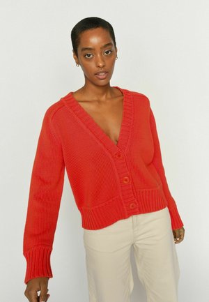 Woman wearing a bright red buttoned knit cardigan with deep V-neck and beige pants against a plain light background.