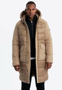Beige Pufferjacke mit Kapuze, gepolstertem Design, zwei Fronttaschen und Reißverschluss. Getragen über einem dunklen Pullover und schwarzen Jeans.
