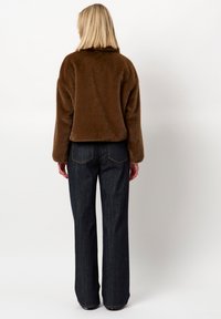 Bruine nep bontjas met een hoge kraag, gecombineerd met rechte donkerdenim jeans. De stof lijkt zacht met een gestructureerde afwerking.