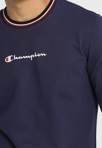 Navyblå sweatshirt i bomullsmix, med rund halsringning och röd- och vitrandig kant samt en vit "Champion"-logotyp på framsidan.