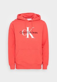 Haut à capuche rouge corail avec poche frontale, cordon de serrage sur la capuche, et logo "Calvin Klein Jeans" blanc et noir sur la poitrine, sur fond blanc.