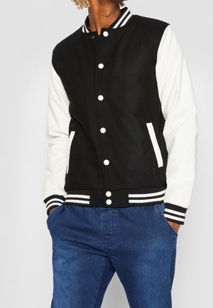 Osoba ubrana w czarno-białą kurtkę klubową z białymi guzikami i niebieskie dżinsy, stojąca z rękami w kieszeniach na jednolitym tle.