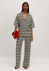 Gestreifter Jumpsuit aus schwarz-weißem Stoff mit kragenförmigem Ausschnitt, kurzen Ärmeln, weitem Bein und strukturiertem Finish. Rote Clutch als Accessoire.