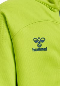 Lys grøn sportstopp lavet af struktureret stof, med blåt "hummel" logo og mønstrede detaljer på skuldrene.