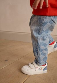 Weiße Sneaker mit Klettverschlüssen, Grafiken von Cartoon-Tieren; hellblaue Akzente. Hellblaue, gemusterte Jeans mit umgeschlagenem Saum. Roter Pullover.