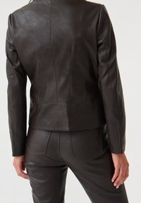 Veste en cuir marron foncé avec un design ajusté, une texture lisse et des coutures sur mesure. Le dos présente une longueur raccourcie et des détails subtils.