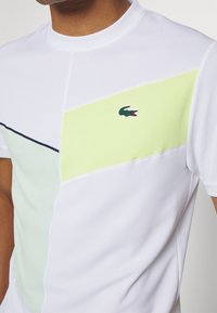 Vit polotröja med gröna och gula geometriska färgblock, med en strukturerad yta och en liten broderad logotyp på bröstet.