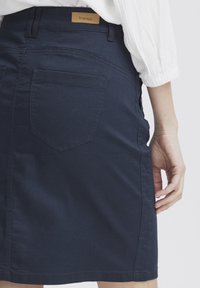 Denim svārki tumši zilos toņos ar šauru piegriezumu. Ar aizmugurējo kabatu un gaiši brūnu etiķeti jostas vietā. Gluda tekstūra un vienkārši šuvju detaļas.