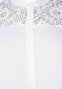 Witte blouse met een gemêleerde voorkant, kantdetails op de schouders en een knoopsluiting met zichtbare kunststof knopen.