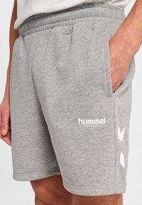 Gros plan sur une personne portant un short de sport Hummel gris avec une main dans la poche et un texte de logo blanc avec des chevrons sur le côté.
