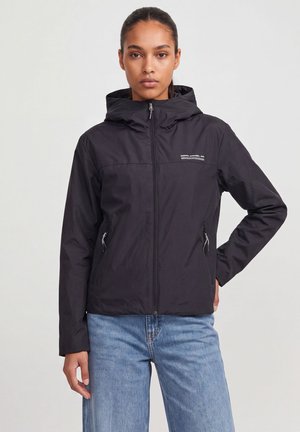 Femme portant une veste noire à capuche avec fermeture éclair et poches zippées, ainsi qu'un jean bleu clair, debout avec une main sur la hanche.