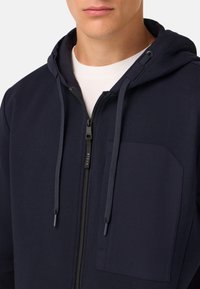 Mornarska zip-up kapuca s kapuco na vezanje, sprednjim žepom in zadrgo. Izdelana iz mehkega, teksturiranega materiala. Ima minimalističen dizajn.